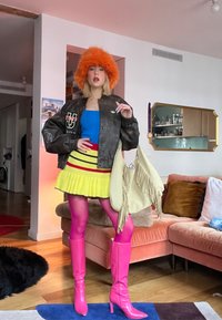 Personne portant un chapeau orange duveteux, une veste bomber foncée, un haut bleu, une jupe jaune, des collants roses et des bottes roses, tenant un grand sac beige à franges à l'intérieur.