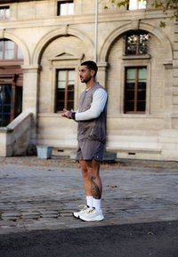 Atlétikai ruhában lévő férfi, aki lábán tetoválás van, áll és csuklóján lévő órát igazítja egy macskaköves járdán, közel egy történelmi épülethez, melynek boltíves ablakai vannak.