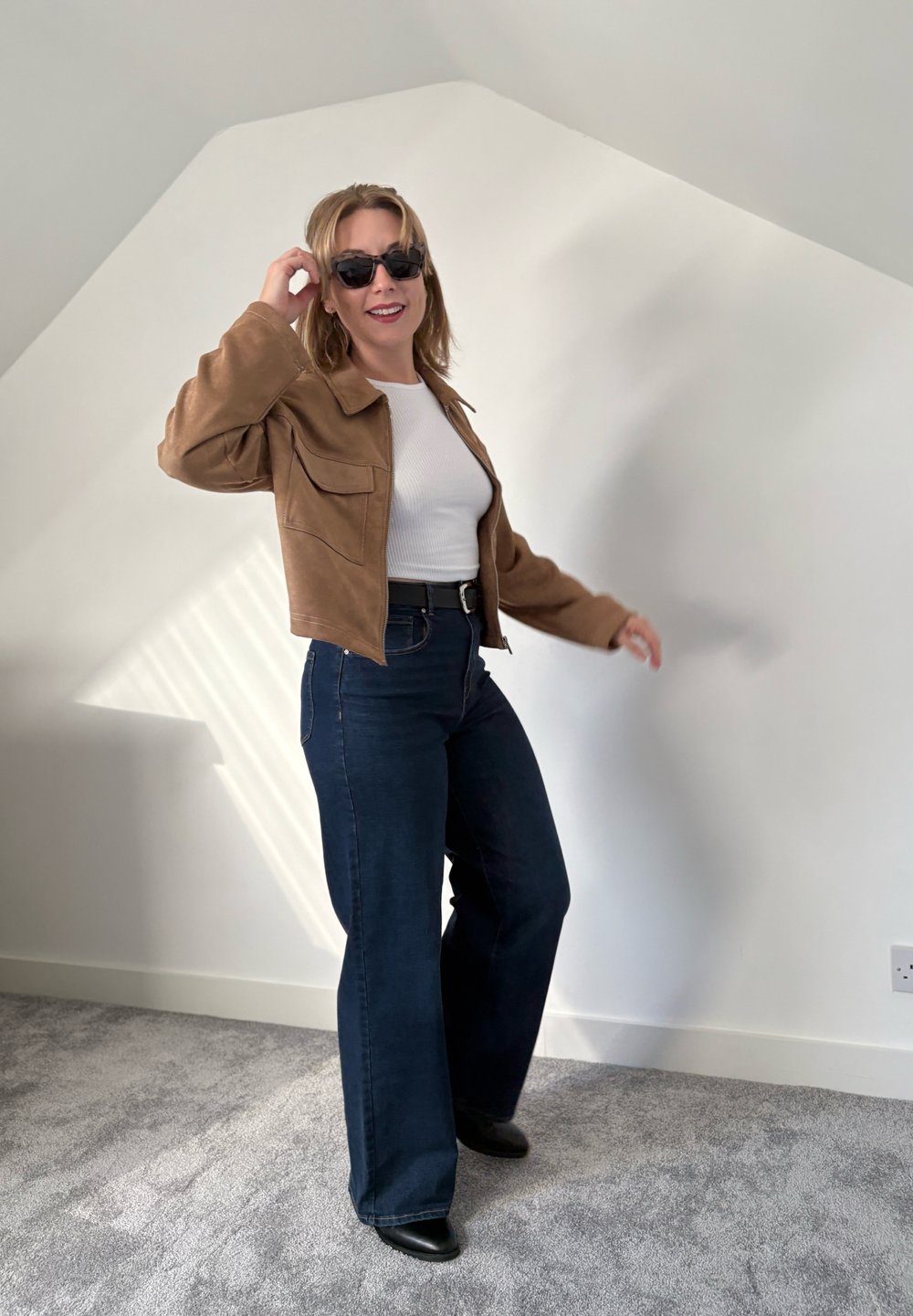 Brown suede jacket, white ribbed top, high-waisted dark blue flared jeans, black ankle boots, and sunglasses. Neutral wall and carpet background.