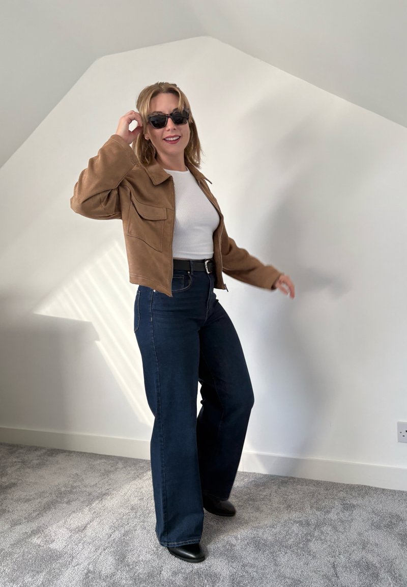 Brown suede jacket, white ribbed top, high-waisted dark blue flared jeans, black ankle boots, and sunglasses. Neutral wall and carpet background.
