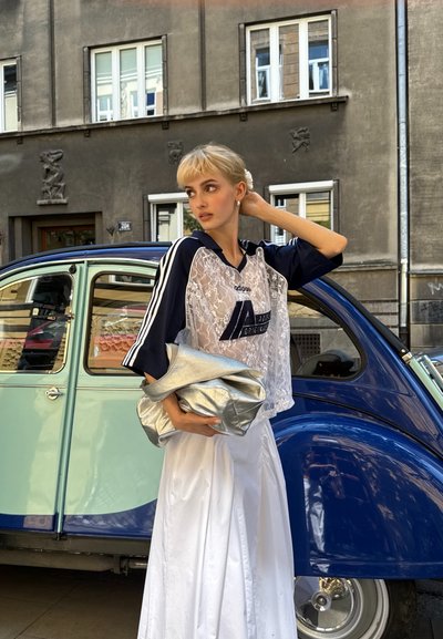 Polo cropped de encaje en azul marino y blanco con tres rayas, combinado con una falda de satén blanca. Sosteniendo un bolso metálico plateado. Coche clásico azul de fondo.