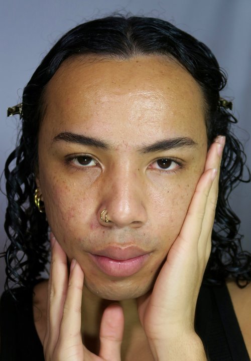 Close-up of a face with curly black hair, orange-red eyeshadow, yellow gradient above eyes, and glossy brown lips. Nose ring visible.
