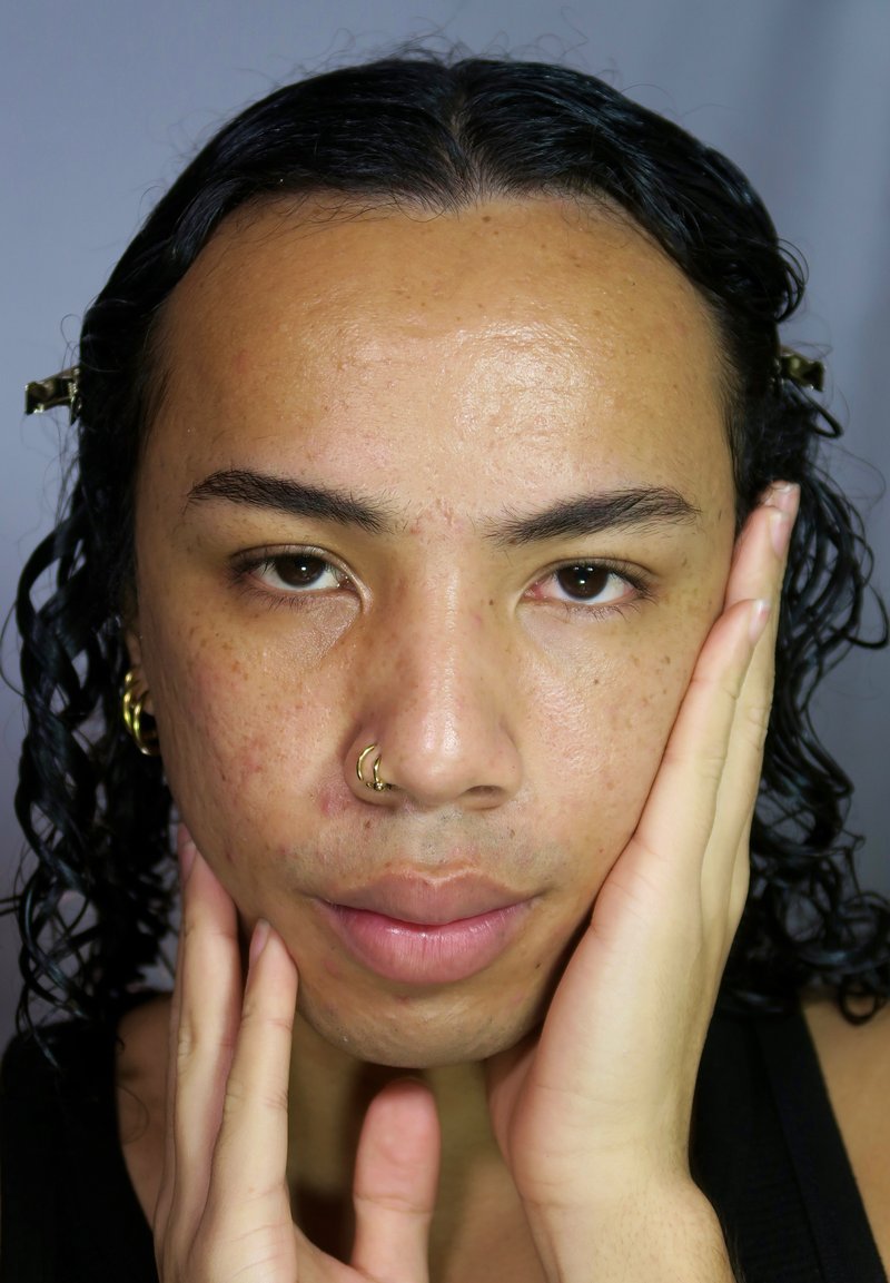 Un retrato en primer plano de una persona con cabello rizado negro, húmedo y de longitud media. Lleva un aro dorado en la nariz y pendientes dorados, y sostiene suavemente su rostro.