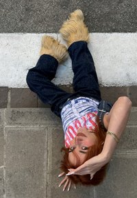 Pehmeät beigenväriset kengät, tummansiniset löysät farkut ja valkoinen grafiikallinen hihaton paita, jossa on punaista ja sinistä tekstiä. Vaaleanruskeat hiukset, kädet kohotettuina poseerauksessa.