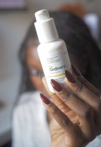 Lotion do opalania w białej plastikowej butelce z pompką. Etykieta zawiera nazwę marki "The Ordinary" oraz szczegóły dotyczące SPF 45. Widoczne ciemnoczerwone paznokcie.