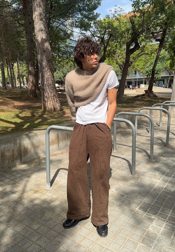 Joven con cabello rizado está de pie en una acera cerca de aparcabicicletas, lleva un chal beige, camisa blanca, pantalones marrones y zapatos negros en un parque.