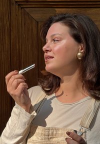 Rouge à lèvres avec un étui argent et crème tenu par une personne portant une chemise beige à manches longues et une salopette claire devant un fond en bois.