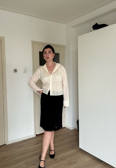 Mujer de pelo oscuro lleva una blusa blanca transparente, falda negra y zapatos negros; está de pie en el interior, sobre un suelo de madera cerca de unas puertas cerradas.
