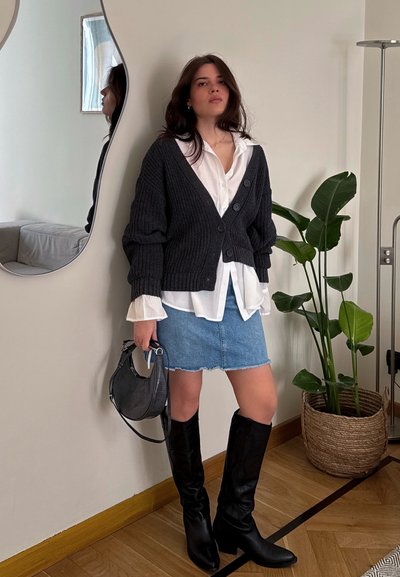 Cárdigan de punto gris oscuro con botones, blusa blanca debajo, falda de mezclilla, botas negras hasta la rodilla, sosteniendo un pequeño bolso negro, en un entorno interior.