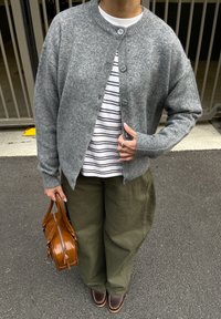 Graue Wollstrickjacke mit Rundhalsausschnitt und Knopfleiste, darüber getragen ein weiß gestreiftes Hemd. Olivgrüne Hosen und eine braune Ledertasche vervollständigen das Outfit.