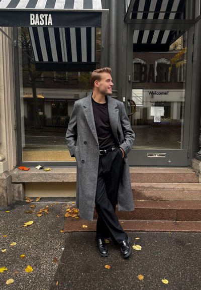 Abrigo gris de doble botonadura, camiseta negra y pantalones. Zapatos negros. De pie en unos escalones con hojas caídas y un toldo a rayas de fondo.