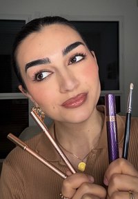 Vier Make-up-Produkte: zwei metallisch roségoldene Stifte, eine lila Wimperntusche mit gewelltem Design und einen dünnen schwarzen Pinsel. Der Hautton ist gebräunt.