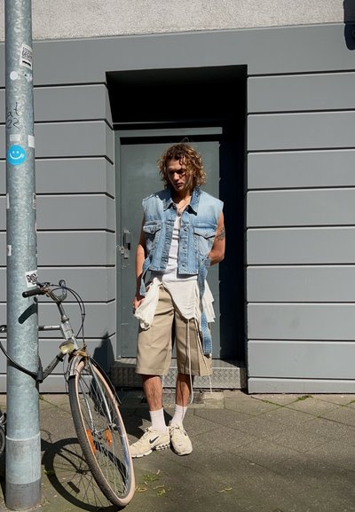 Chaleco de mezclilla sobre camisa blanca, pantalones cortos beige y zapatillas crema. De pie junto a una bicicleta contra una pared gris con una puerta detrás.