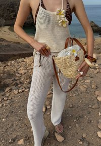 Top din croșetat și pantaloni în off-white. Geantă din paie împletită cu mânere din piele maro și accent de floare albă. Sandale plate cu curea curbă.