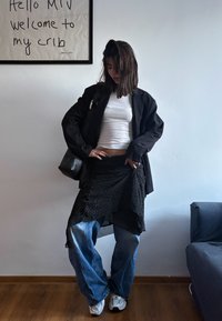 Veste noire surdimensionnée, haut blanc ajusté, jupe noire à pois superposée, jean bleu ample et baskets blanches dans un intérieur minimaliste.