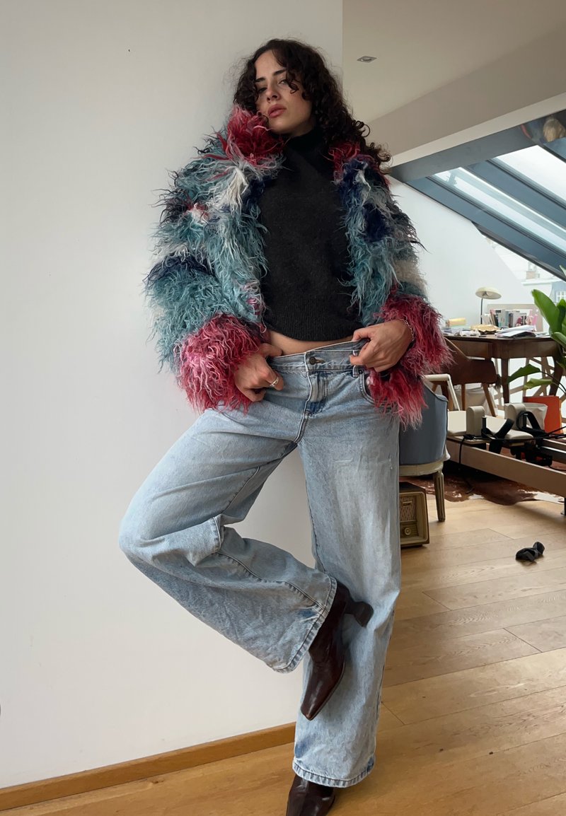 Nepbont multikleurige jas met tinten roze, blauw en wit, gecombineerd met een zwarte gebreide top en lichtblauwe jeans met wijde pijpen. Bruine enkellaarsjes.