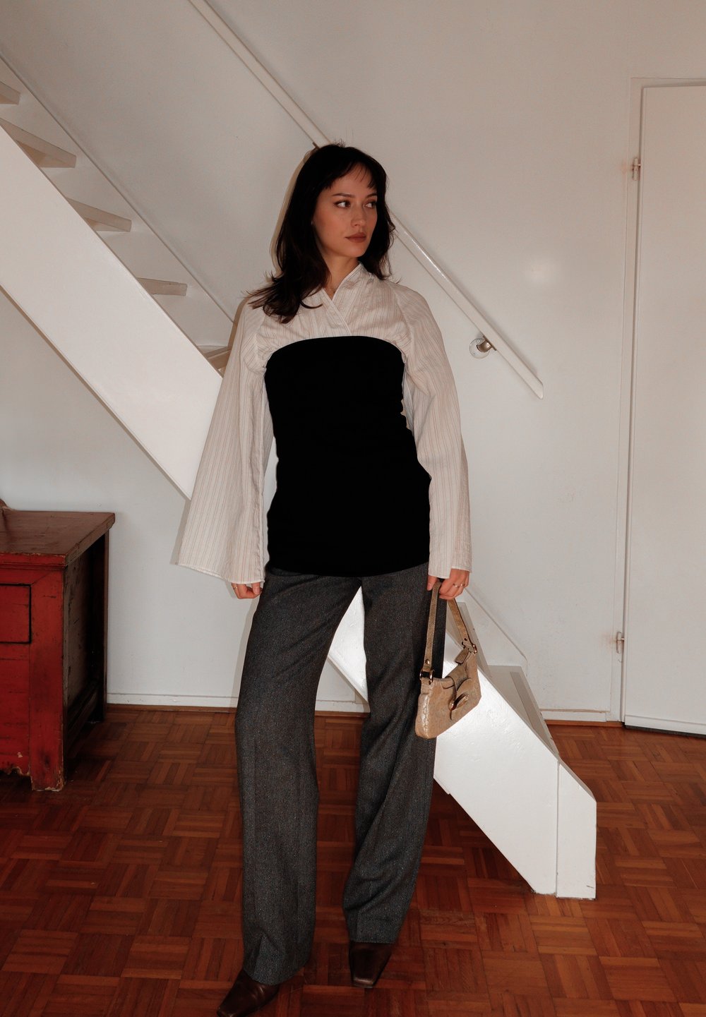 Top em camadas em preto e branco com mangas flare, calças cinzentas texturizadas, sapatos de salto castanhos e uma pequena mala bege. Fundo simples.