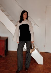 Top em camadas em preto e branco com mangas flare, calças cinzentas texturizadas, sapatos de salto castanhos e uma pequena mala bege. Fundo simples.
