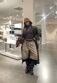 Ruskea nahkainen pitkä takki tumman ribatun paidan päällä, beige korkeavyötäröiset shortsit ja mustat polvimittaiset saappaat. Beige kuvioitu bucket-hattu, hopeiset korut.