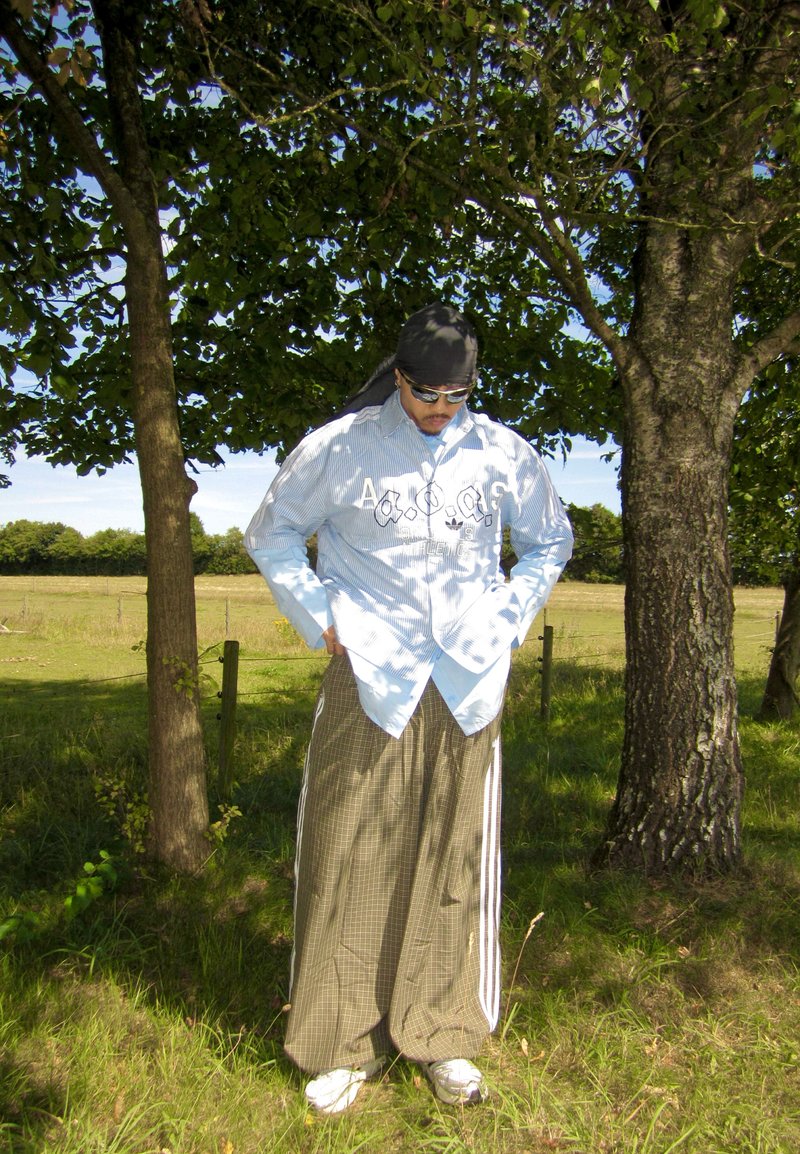 Homme portant des lunettes de soleil, un foulard noir, une chemise rayée et un pantalon large marron à carreaux, debout les mains dans les poches sous un arbre dans un champ ensoleillé.