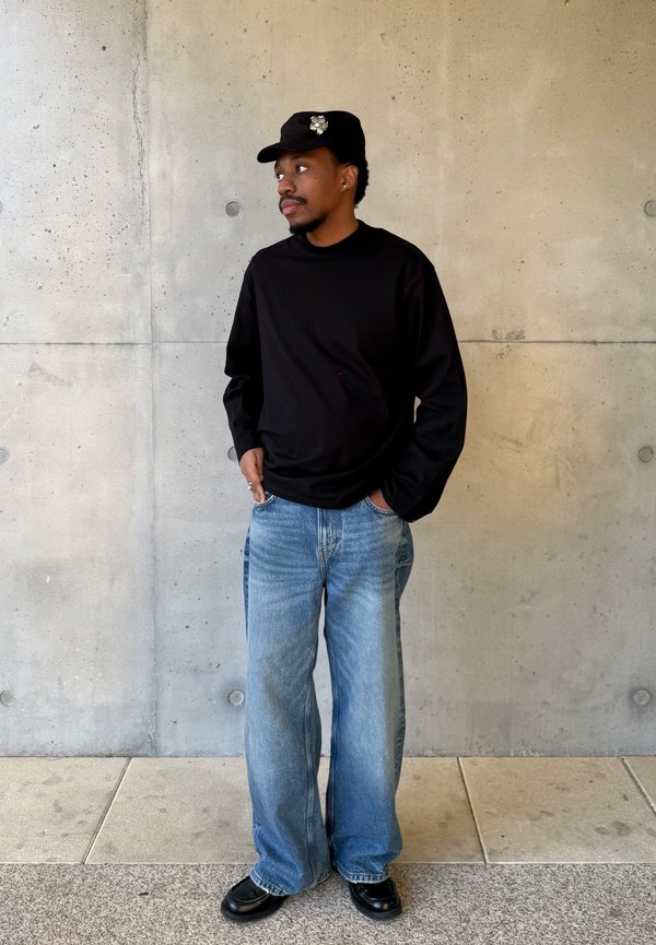 Mężczyzna w czarnej koszuli z długim rękawem, szerokich niebieskich dżinsach, czarnych butach i czarnej czapce z ozdobną przypinką, stojący przy betonowej ścianie.