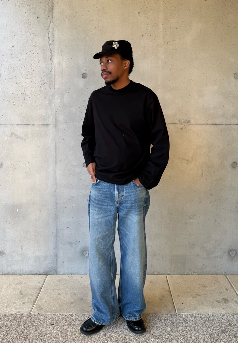 Uomo con camicia nera a maniche lunghe, jeans blu a gamba larga, scarpe nere e cappellino nero con spilla decorativa, in piedi contro un muro di cemento.