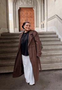 Rudos spalvos tranšėjos paltas dėvimas ant juodos palaidinės ir baltų plačių kelnių, stovint ant akmeninių laiptų, fone matyti medinės durys.