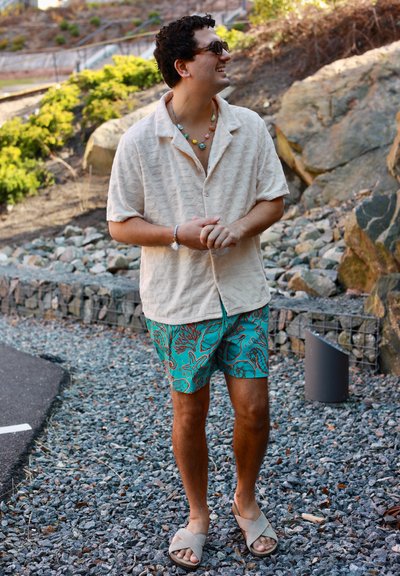 Hombre con camisa beige de manga corta, pantalones cortos turquesa con estampado, sandalias y collar de cuentas, de pie en un camino de grava al aire libre cerca de unas rocas.