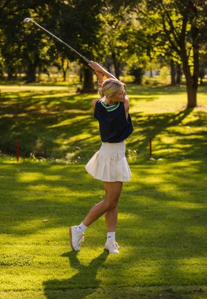 Mujer con un chaleco azul marino y una falda plisada blanca balanceando un palo de golf en un campo de hierba iluminado por el sol con árboles al fondo.
