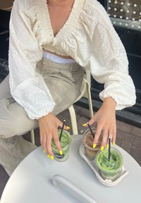 White textured blouse with puff sleeves, light beige trousers, and bright yellow nails holding iced drinks on a round table with a beige holder.
