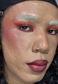 Nahaufnahme eines Gesichts mit strukturierter Haut, blassen Augenbrauen, kräftig dunklen Lippen in Burgunderrot und schimmerndem orangefarbenem Lidschatten. Mit einer goldenen Septumpiercing.