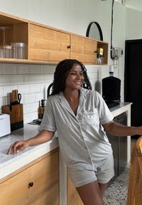 Femme souriante appuyée sur le comptoir de la cuisine, portant un haut gris à manches courtes boutonné et un short assorti dans une cuisine moderne.