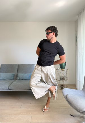Hombre con gafas de sol, camisa negra y pantalones anchos beige, de pie sobre un pie con las manos detrás de la espalda en una sala de estar moderna.