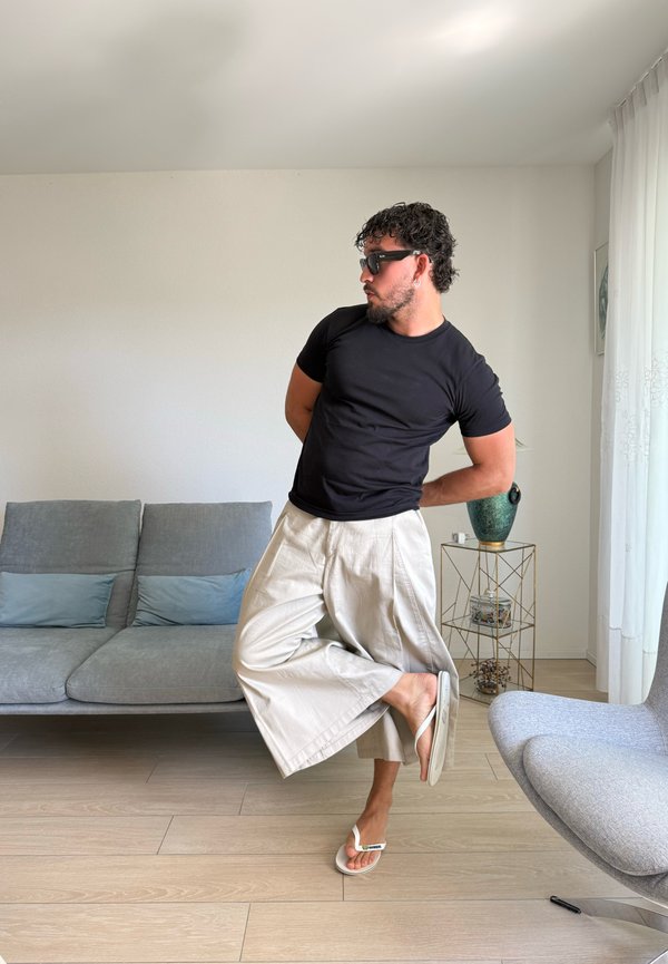 Hombre con gafas de sol, camisa negra y pantalones anchos beige, de pie sobre un pie con las manos detrás de la espalda en una sala de estar moderna.