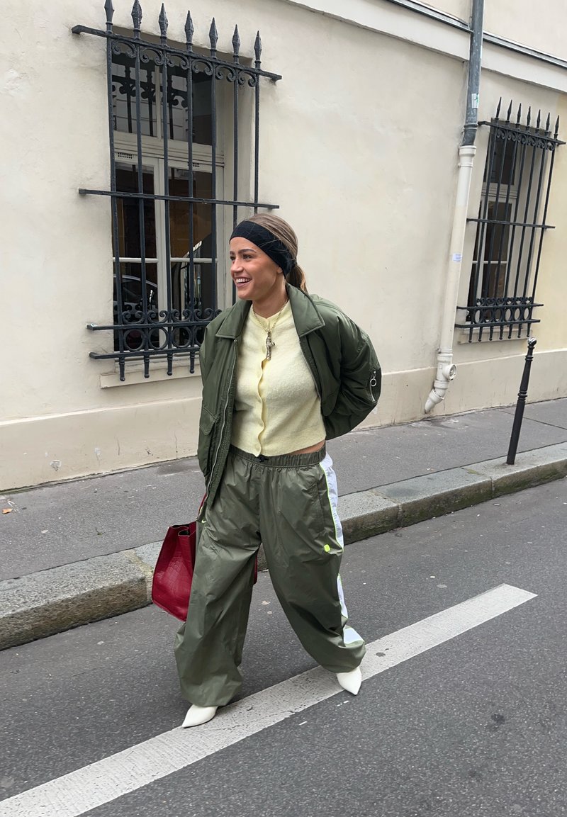Femme souriante marchant dans la rue, portant une veste et un pantalon verts, un haut jaune, des chaussures blanches, un bandeau noir et un sac à main rouge.