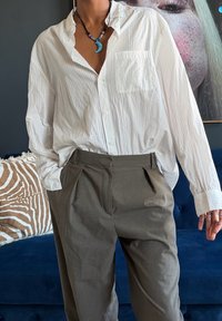 Personne portant une chemise blanche à boutons ample, un pantalon gris et un collier bleu en forme de croissant de lune, debout devant un canapé bleu avec un coussin à motifs.