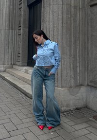 Chemise crop bleu clair à boutons avec des accents en noeud, associée à un jean large bleu délavé et des escarpins rouges à bout pointu.