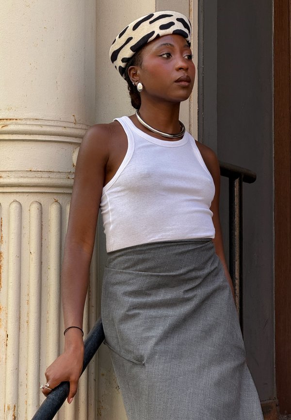 Mujer con camiseta de tirantes blanca, falda gris y boina de patrón en blanco y negro apoyada en una barandilla negra junto a una columna.