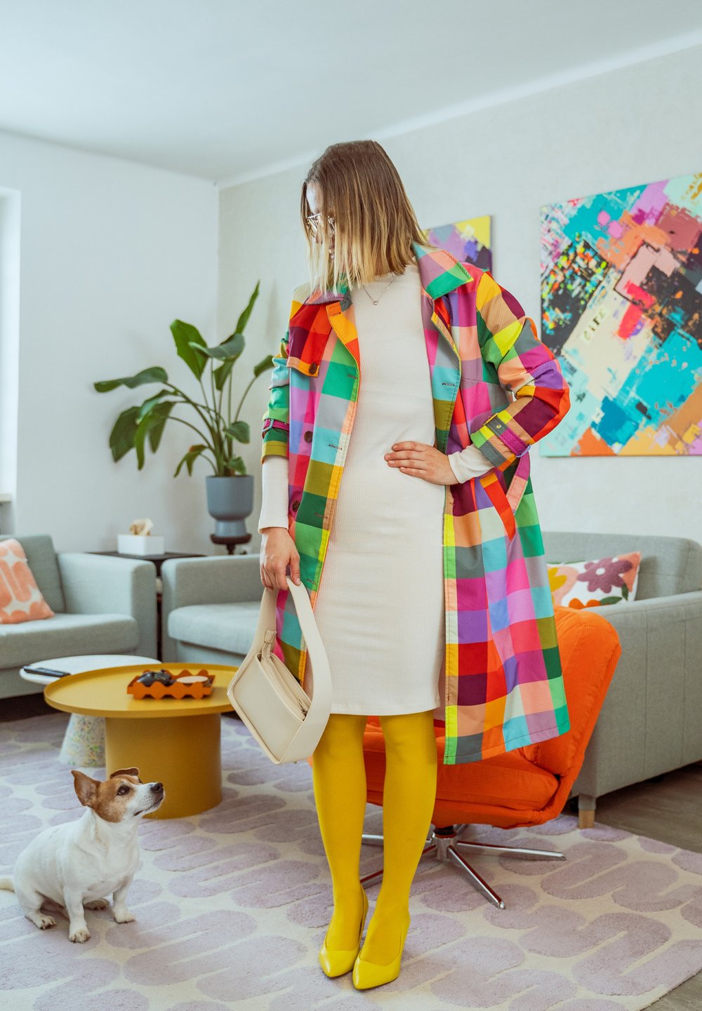 Femme portant un manteau à carreaux colorés, une robe blanche, des collants jaunes et des talons, tenant un sac beige, debout dans un salon avec un petit chien à proximité.