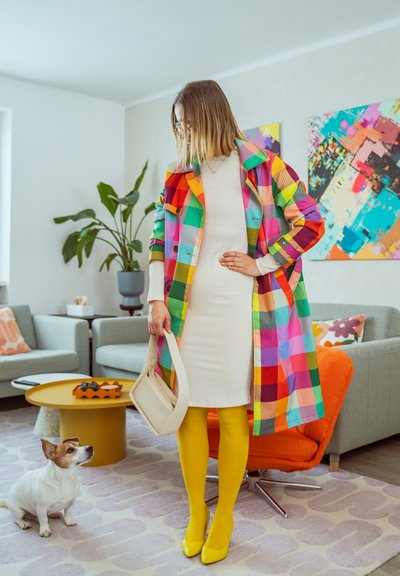 Mujer con abrigo de cuadros coloridos, vestido blanco, medias amarillas y tacones, sosteniendo un bolso beige, de pie en una sala de estar con un perro pequeño cerca.