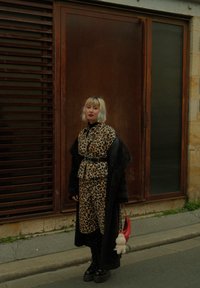 Plettet leopardmønstret frakk og kjole med belte, svarte knærne høye støvler, og en rød veske med en fluffy charm, satt mot en teksturert brun bakgrunn.