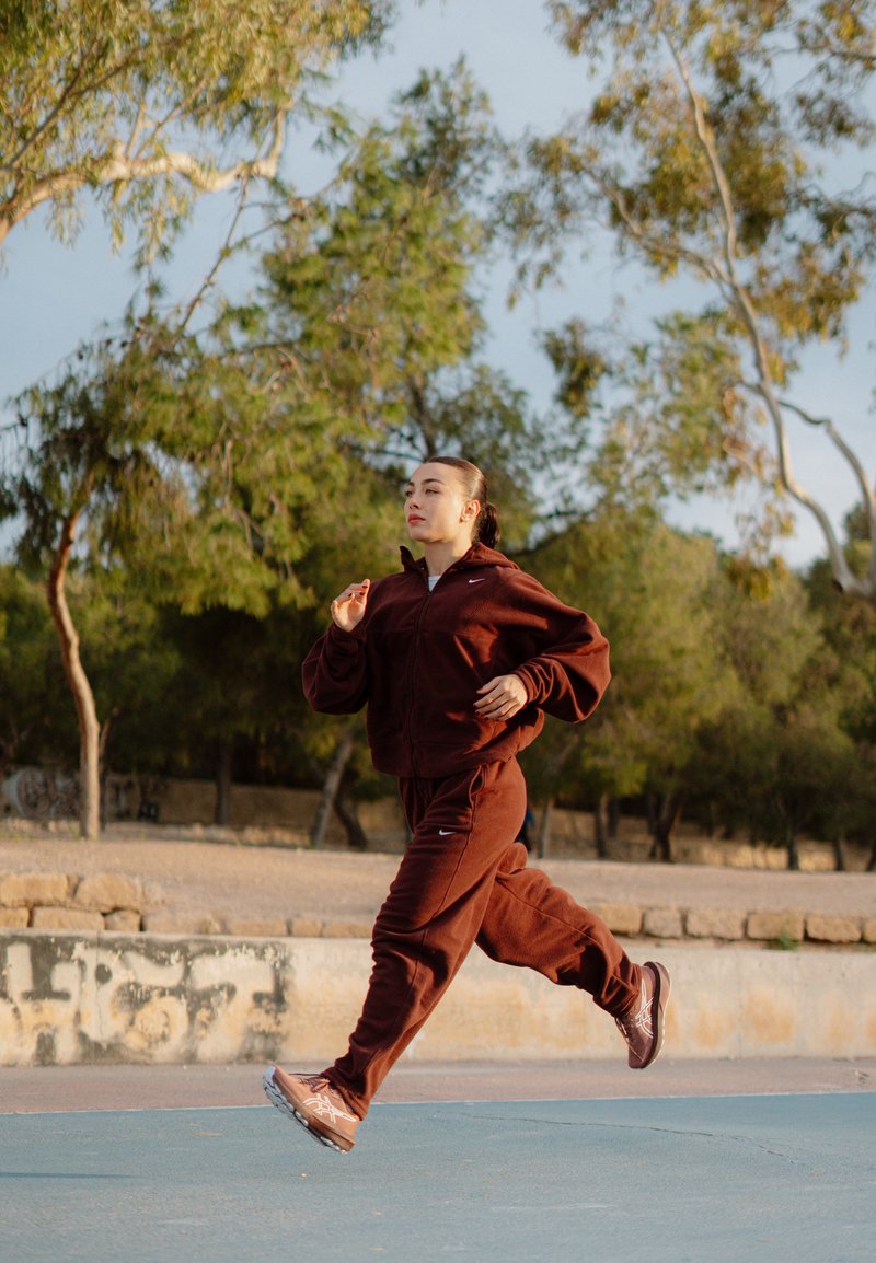 Γυναίκα με καφέ αθλητική ένδυση τρέχει έξω σε στρωμένο μονοπάτι με δέντρα και πέτρινο τοίχο στο παρασκήνιο.