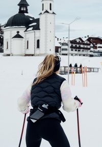 Γυναίκα με χειμερινά ρούχα κρατώντας μπαστούνια σκι στέκεται στο χιόνι και κοιτάζει μια λευκή εκκλησία με καμπαναριό σε ένα χιονισμένο χωριό.