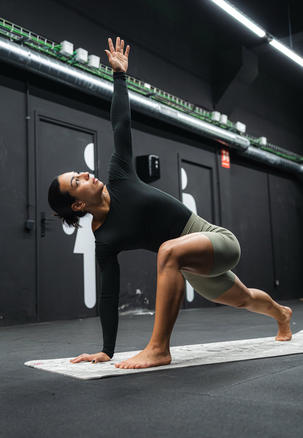 En person iført en sort langærmet top og lysegrønne shorts står i en skridtlunge med den ene arm hævet på en yogamåtte. Baggrunden er mørk.