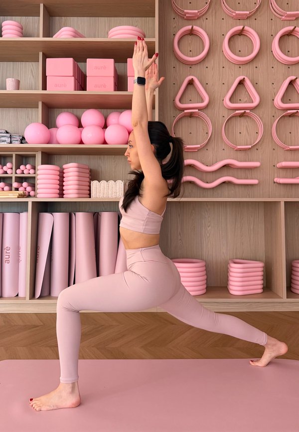 Rózsaszín aktív ruhában lévő nő magas kitöréses jóga pózt végez egy rózsaszín matracon, mögötte fapolcokon rózsaszín fitneszeszközökkel.