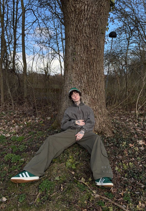 Persoon met een groene pet, een grijze jas en groene sneakers, zittend met gespreide benen tegen een grote boom in een bosrijk gebied.