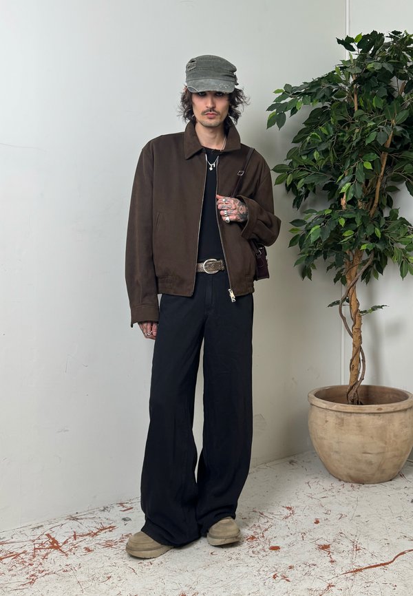 Personne portant une veste brun foncé, une chemise noire, un pantalon large noir, une casquette grise, debout à côté d'une plante en pot à feuilles contre un mur uni.