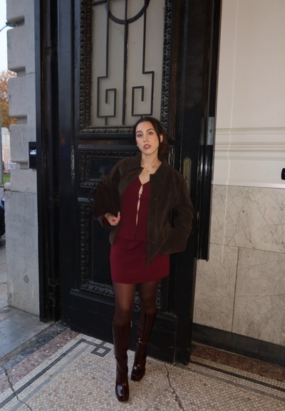 Chaqueta de ante marrón sobre un vestido burdeos, combinada con botas altas de charol color ciruela. De pie frente a una puerta negra ornamentada y una pared de mármol.