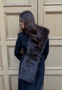 Femme aux longs cheveux noirs portant un manteau noir et un col en fourrure brun foncé maintenu par une ceinture noire, debout contre une porte en bois.