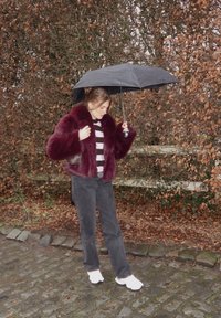 Femme en manteau de fourrure bordeaux et pull rayé tenant un parapluie noir sur un chemin pavé avec un fond de feuilles d'automne brunes.